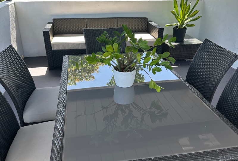 A spacious covered outdoor patio with a dark wicker dining table, glass tabletop, grey cushioned chairs, a potted ZZ plant, and a matching outdoor lounge sofa in the background.