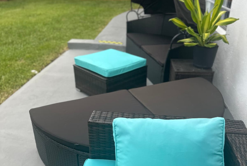 Modern dark wicker patio seating featuring vibrant turquoise-blue seat cushions and pillows, a central coffee table, and a canopied daybed set on a light gray concrete surface in a residential backyard.