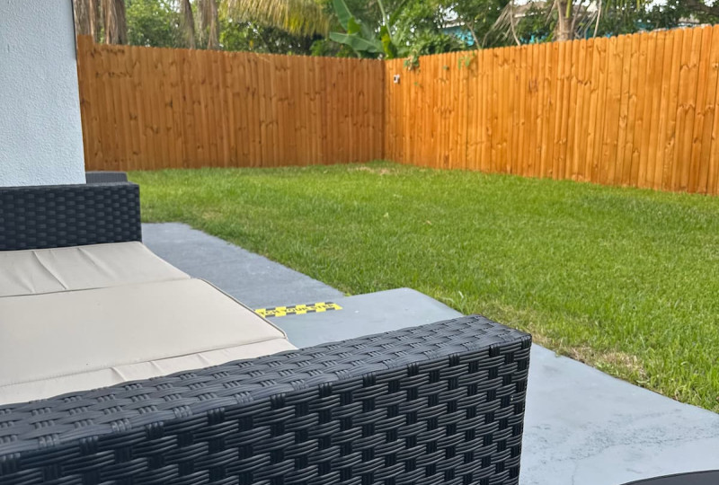 Black rattan patio sofa looking out at a grassy backyard with a tall wooden fence and palm trees