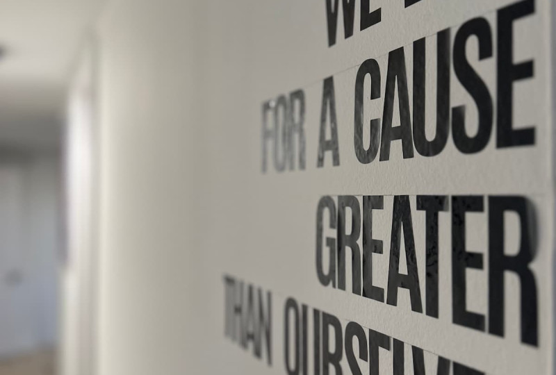 Angled side shot of black block letters on a white wall spelling out We Live For A Cause Greater Than Ourselves in an indoor hallway