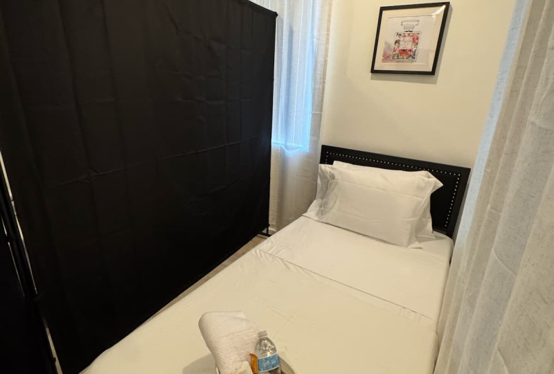 A single bed made with white sheets, separated by a black privacy screen divider next to a window with sheer white curtains.
