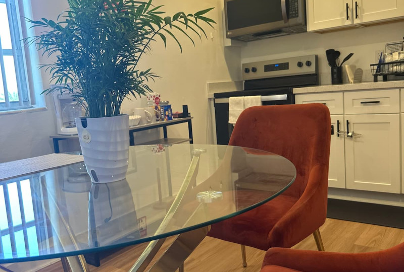 A round glass-top dining table with gold metallic legs and rust-orange velvet upholstered chairs, featuring a potted palm centerpiece, set adjacent to a modern white kitchen with stainless steel appliances.
