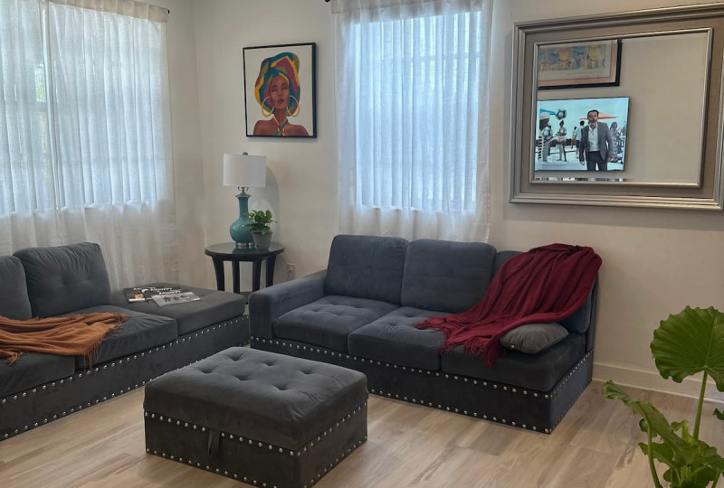 Dark gray sofas and a matching ottoman in a bright living room with light vinyl plank flooring and large windows with sheer curtains.